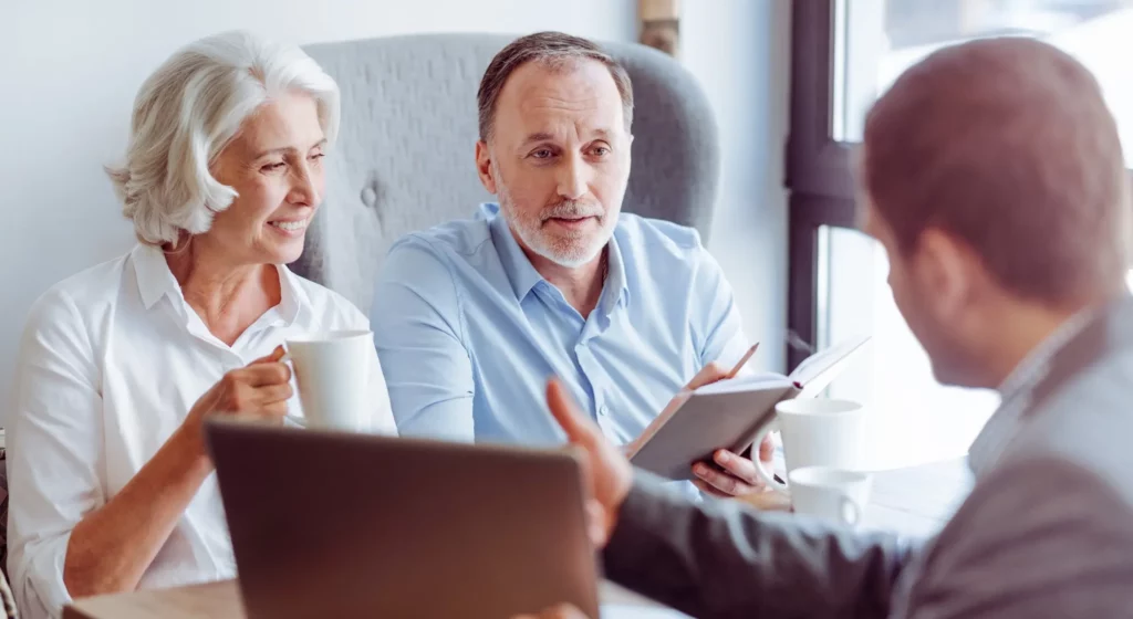 Couple senior souriant échangeant autour d'une table avec un conseiller lors d'un entretien de prévoyance obsèques.